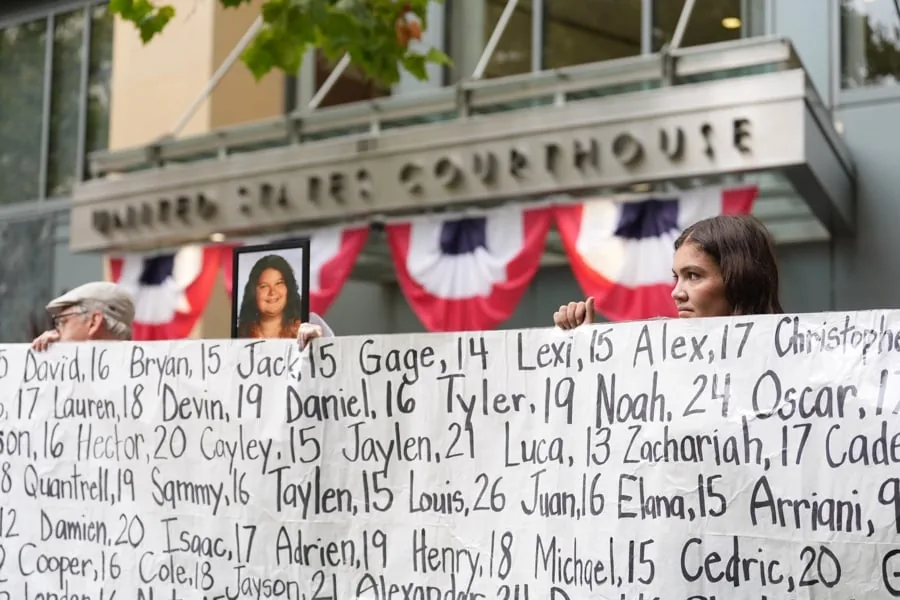 Manifestantes con pancarta de nombres y edades frente a un edificio que dice 'United States Courthouse', decorado con banderines patrióticos. Una mujer sostiene una foto.