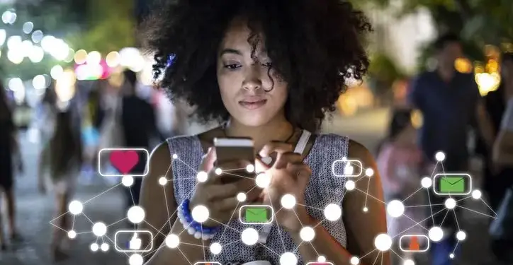 Mujer de cabello rizado usa un teléfono móvil en la calle rodeada de gráficos digitales de redes sociales.