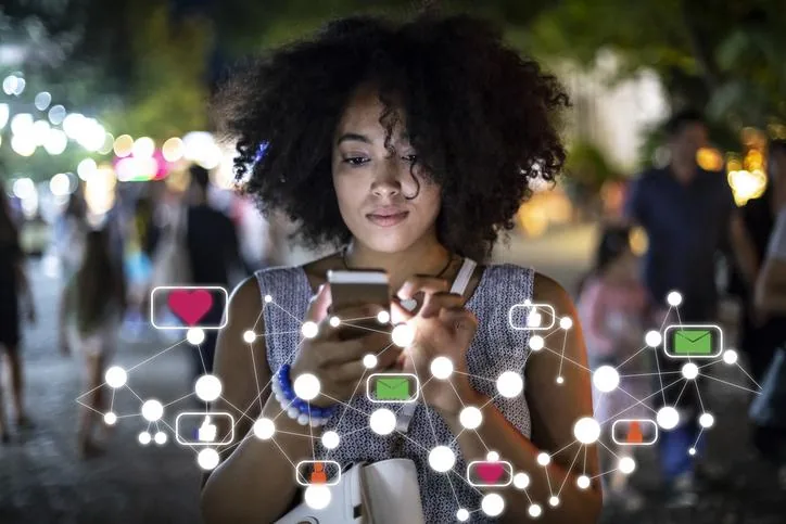 Mujer de cabello rizado usa un teléfono móvil en la calle rodeada de gráficos digitales de redes sociales.