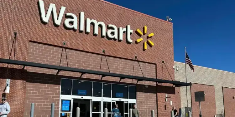 Fachada de una tienda Walmart de ladrillo rojo con su logo blanco y amarillo. Personas entran y salen por puertas automáticas. Bandera de EE. UU. ondea.