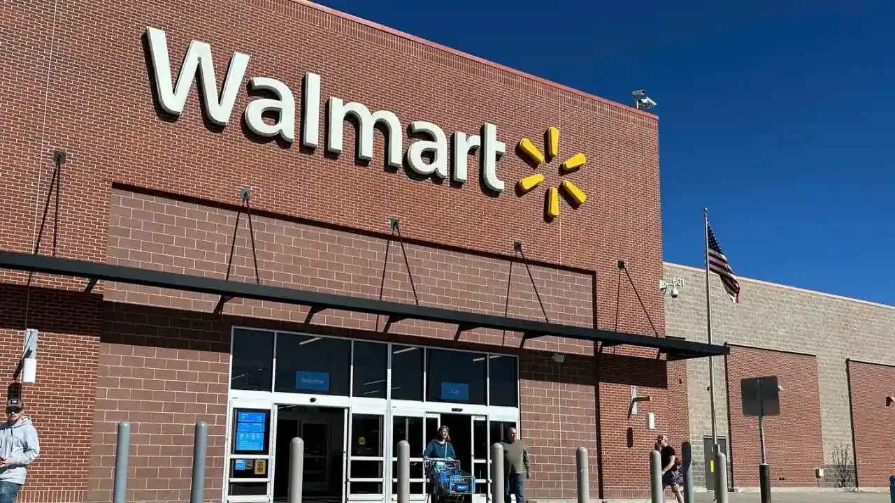 Fachada de una tienda Walmart de ladrillo rojo con su logo blanco y amarillo. Personas entran y salen por puertas automáticas. Bandera de EE. UU. ondea.