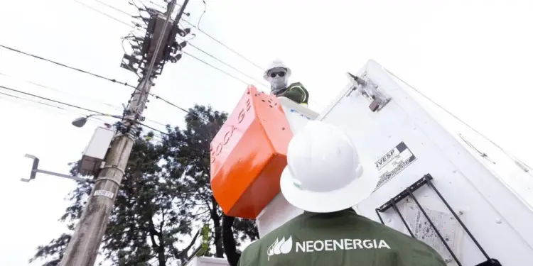 Dos técnicos con cascos de seguridad realizan mantenimiento en un poste de energía eléctrica con transformador y cables, usando una plataforma elevadora. Uno lleva uniforme de NEOENERGIA.