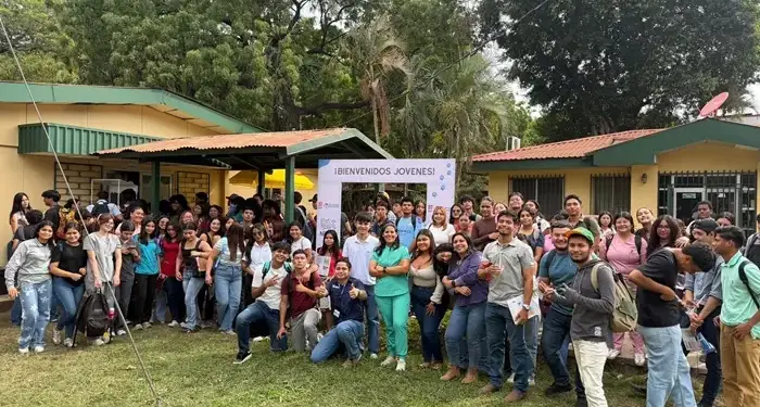 Numeroso grupo de jóvenes y adultos posando juntos al aire libre frente a edificios de una planta rodeados de árboles.
