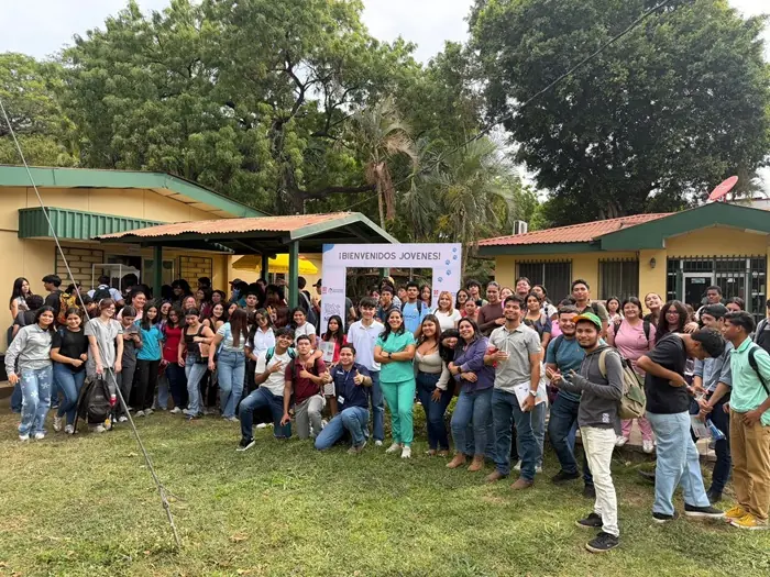 Numeroso grupo de jóvenes y adultos posando juntos al aire libre frente a edificios de una planta rodeados de árboles.