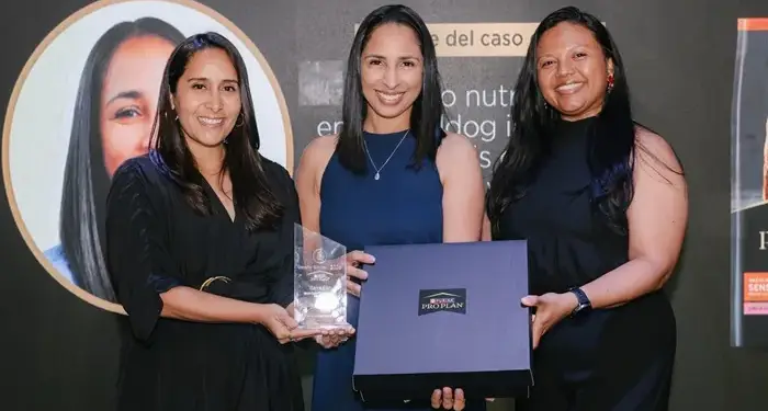 Tres mujeres profesionales sonriendo mientras sostienen un trofeo y una caja con el logotipo de Purina Pro Plan.