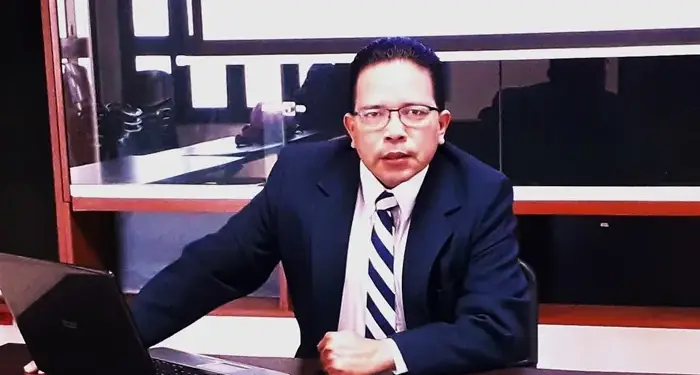 Hombre profesional con traje azul marino y corbata a rayas, sentado en un escritorio de oficina con un portátil abierto. Su expresión es seria.