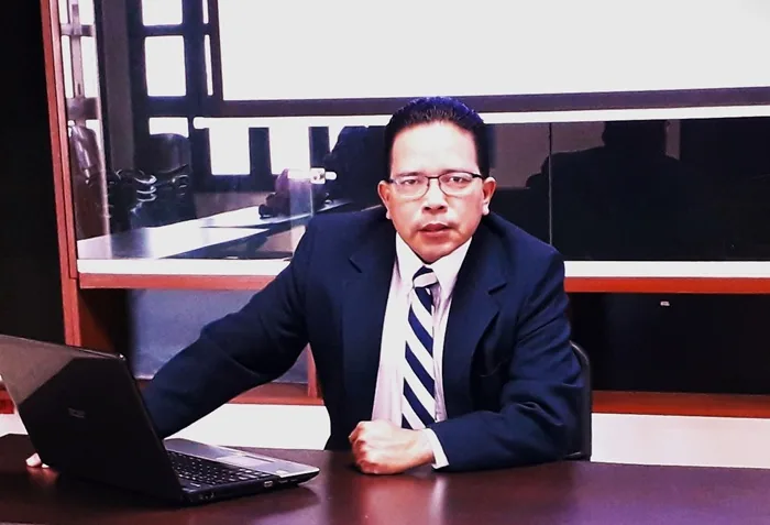 Hombre profesional con traje azul marino y corbata a rayas, sentado en un escritorio de oficina con un portátil abierto. Su expresión es seria.
