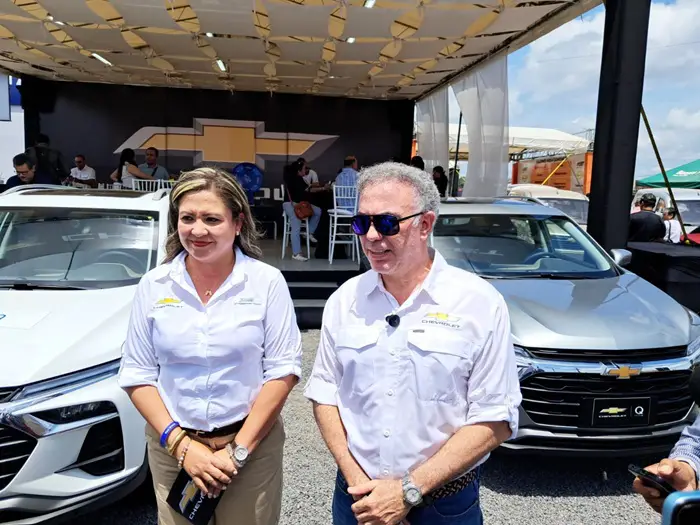 Dos profesionales, hombre y mujer, frente a SUVs Chevrolet blancos y plateados en evento automotriz. Fondo con logo Chevrolet.