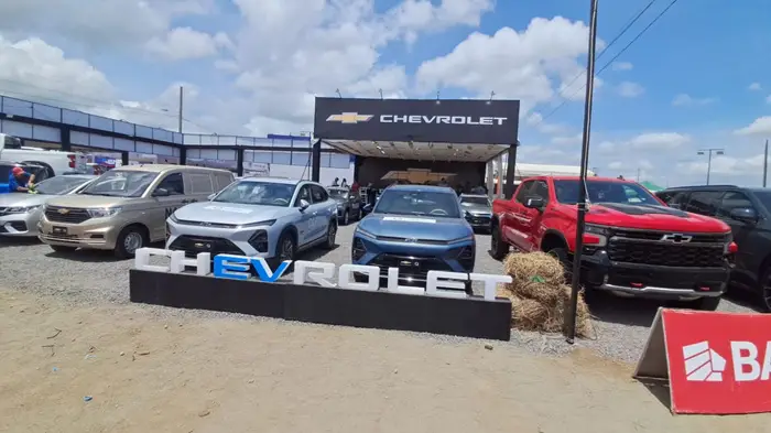 Exhibición al aire libre de vehículos Chevrolet, con una van dorada, dos SUVs azules/plateadas y una camioneta roja, bajo un gran cartel de la marca.
