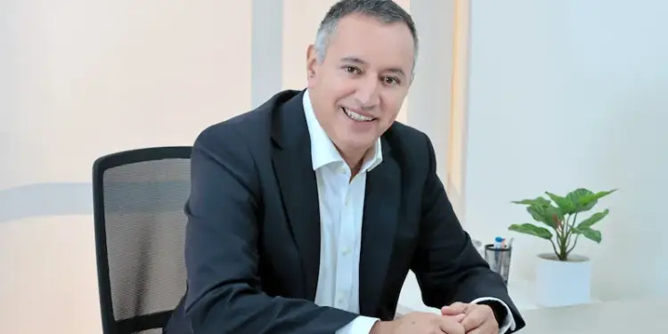 Hombre de mediana edad, cabello gris, sonriendo, vestido con traje oscuro y camisa blanca, sentado en un escritorio de oficina moderno con una planta verde.