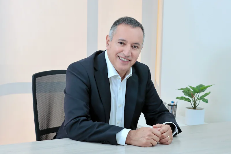 Hombre de mediana edad, cabello gris, sonriendo, vestido con traje oscuro y camisa blanca, sentado en un escritorio de oficina moderno con una planta verde.