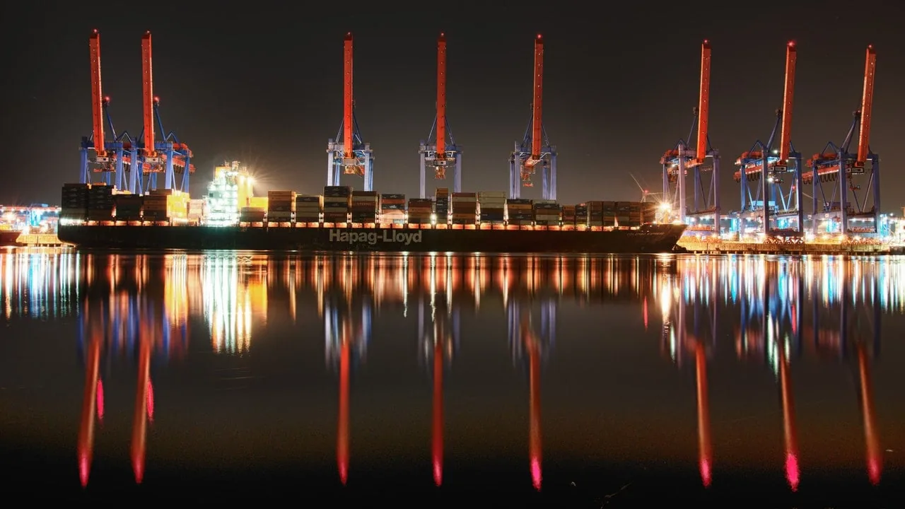 Gran buque de carga Hapag-Lloyd con contenedores en un muelle iluminado por la noche, rodeado de altas grúas y reflejos vibrantes.