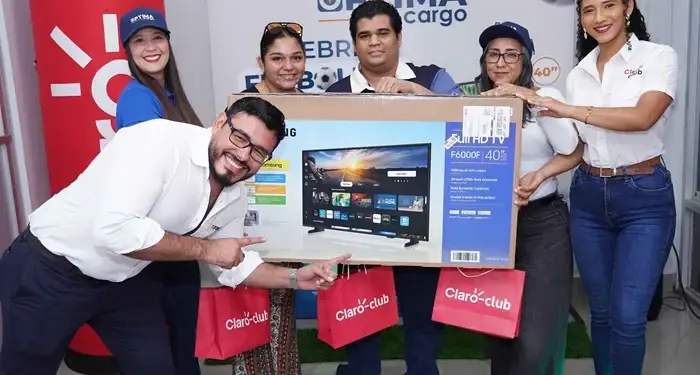 Grupo de cinco personas sonriendo mientras sostienen una caja grande con un televisor y bolsas de regalo rojas en una oficina.
