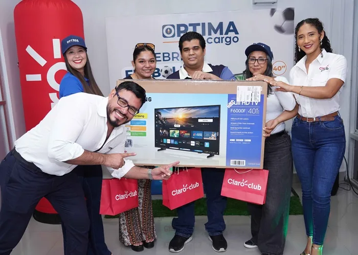 Grupo de cinco personas sonriendo mientras sostienen una caja grande con un televisor y bolsas de regalo rojas en una oficina.