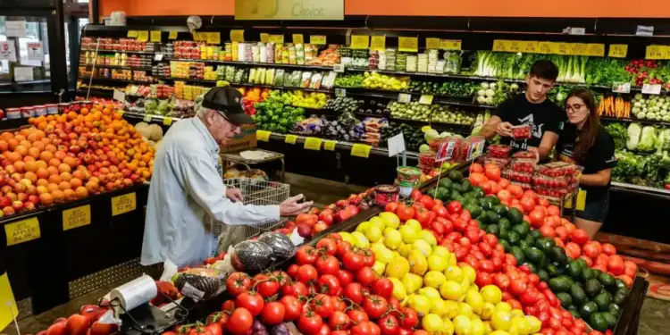 Cliente de la tercera edad comprando frutas y verduras en el pasillo de un supermercado junto a empleados trabajando.