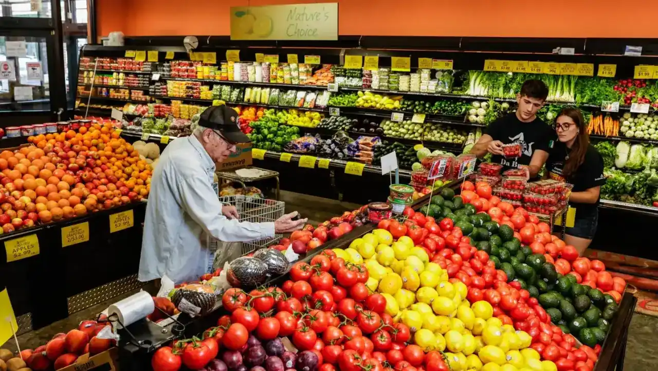 Cliente de la tercera edad comprando frutas y verduras en el pasillo de un supermercado junto a empleados trabajando.