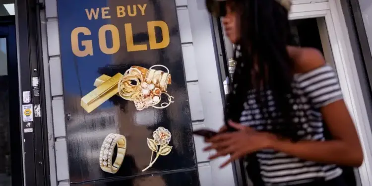 Una mujer caminando frente a un letrero negro que dice WE BUY GOLD con imágenes de lingotes y joyas de oro.