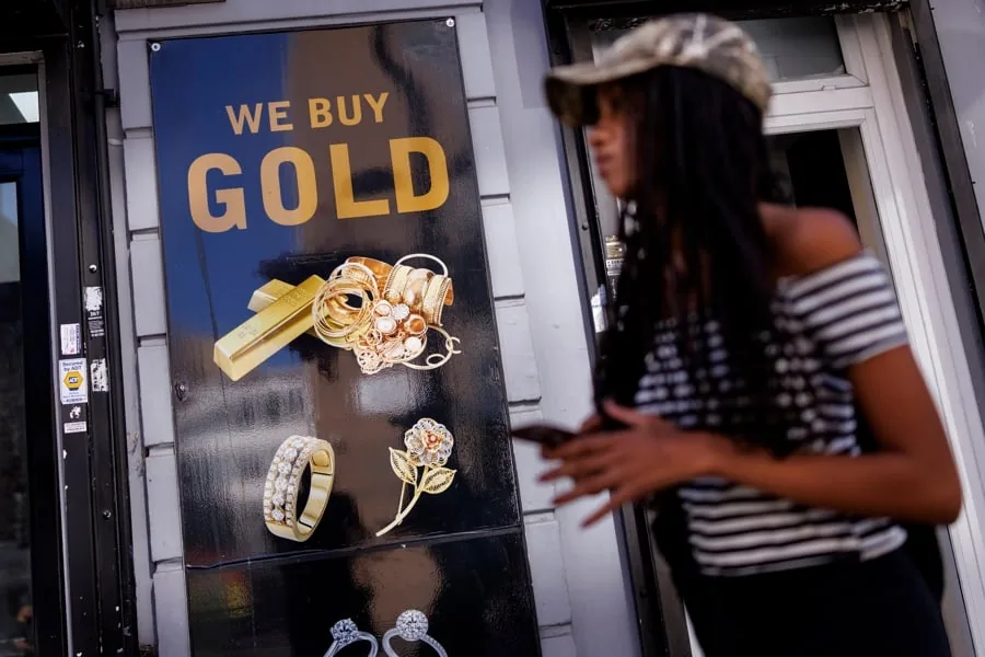 Una mujer caminando frente a un letrero negro que dice WE BUY GOLD con imágenes de lingotes y joyas de oro.