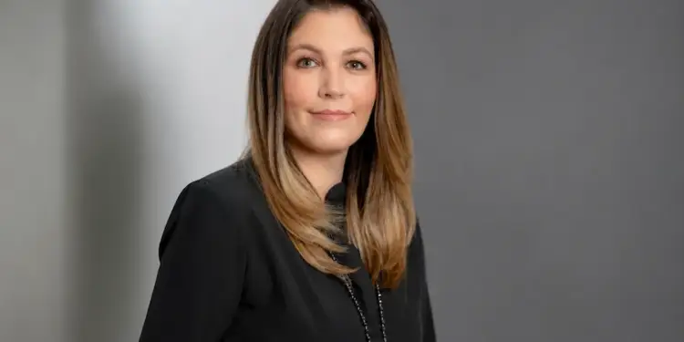 Retrato de una mujer profesional de mediana edad con cabello castaño claro, vestida con una blusa negra y un collar, mirando a la cámara con una sonrisa sutil. Fondo gris.