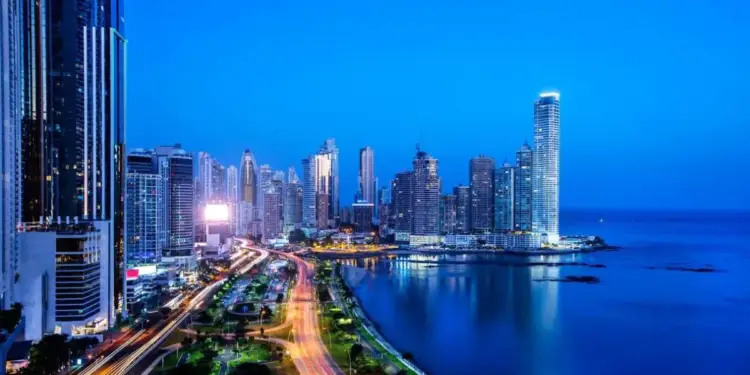 Vista nocturna de un distrito financiero con rascacielos modernos, una carretera con tráfico y la costa marítima iluminada.