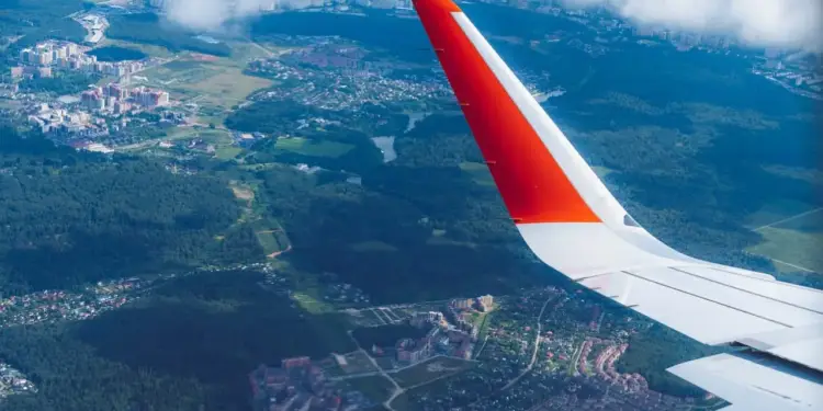 Vista aérea desde la ventanilla de un avión mostrando el ala con punta naranja y un paisaje urbano y natural abajo.