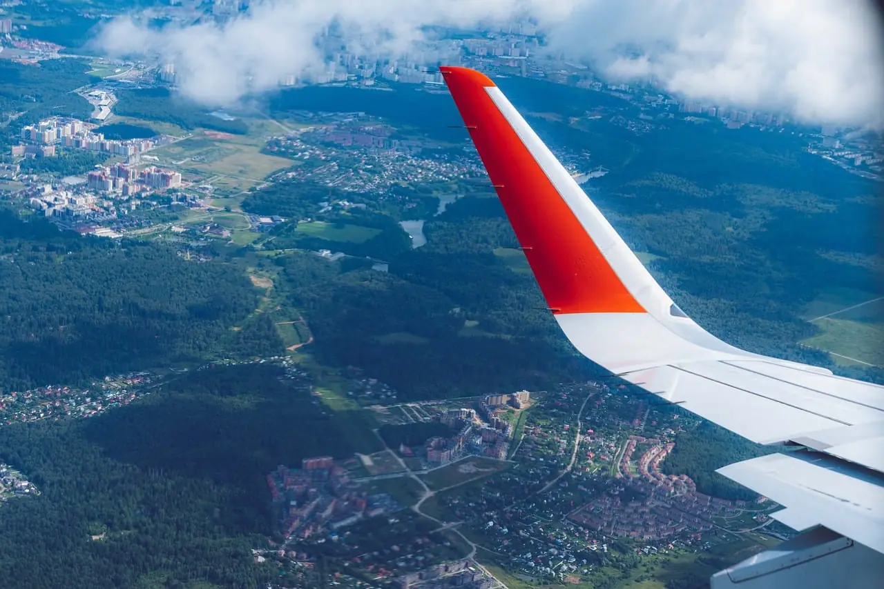 Vista aérea desde la ventanilla de un avión mostrando el ala con punta naranja y un paisaje urbano y natural abajo.