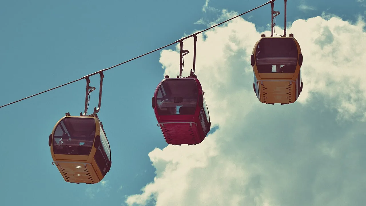 Tres cabinas de teleférico de colores amarillo y rojo suspendidas de un cable contra un fondo de cielo azul y nubes blancas.