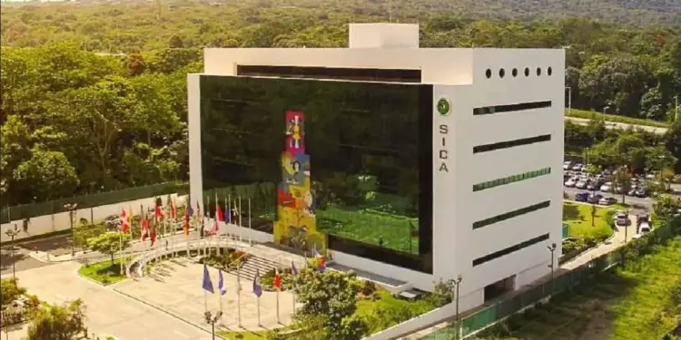 Edificio moderno de SICA con fachada blanca y ventanas reflectantes, rodeado de banderas nacionales, árboles y un estacionamiento con autos.