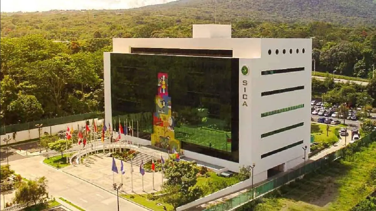 Edificio moderno de SICA con fachada blanca y ventanas reflectantes, rodeado de banderas nacionales, árboles y un estacionamiento con autos.