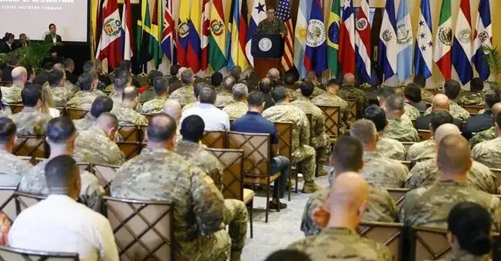 Audiencia de militares y civiles escuchando una conferencia frente a un estrado con banderas.