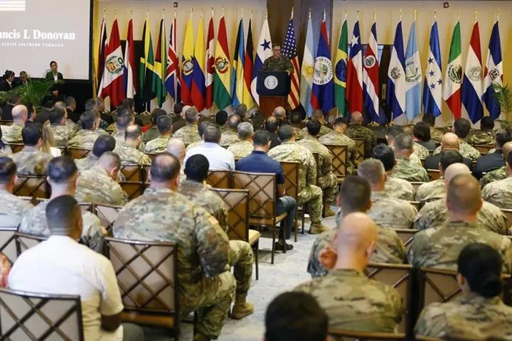 Audiencia de militares y civiles escuchando una conferencia frente a un estrado con banderas.