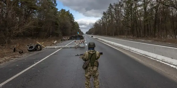 Soldado armado vigilando una carretera bloqueada con barricadas y neumáticos en una zona boscosa.