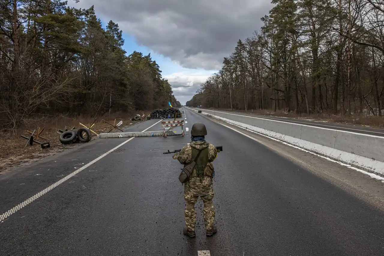 Soldado armado vigilando una carretera bloqueada con barricadas y neumáticos en una zona boscosa.