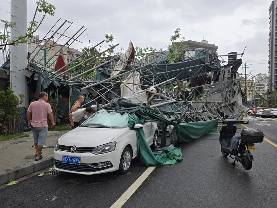 Vista de una estructura de andamios y lonas verdes derrumbada sobre un vehículo blanco en una calle urbana, con tres personas evaluando los daños.