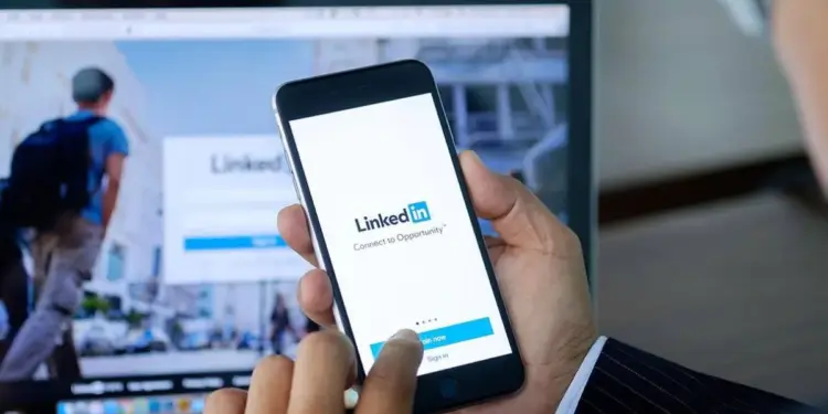 Una persona con traje sostiene un smartphone mostrando la app de LinkedIn, con el logo y 'Connect to Opportunity'. Un portátil en el fondo también muestra la web de LinkedIn.