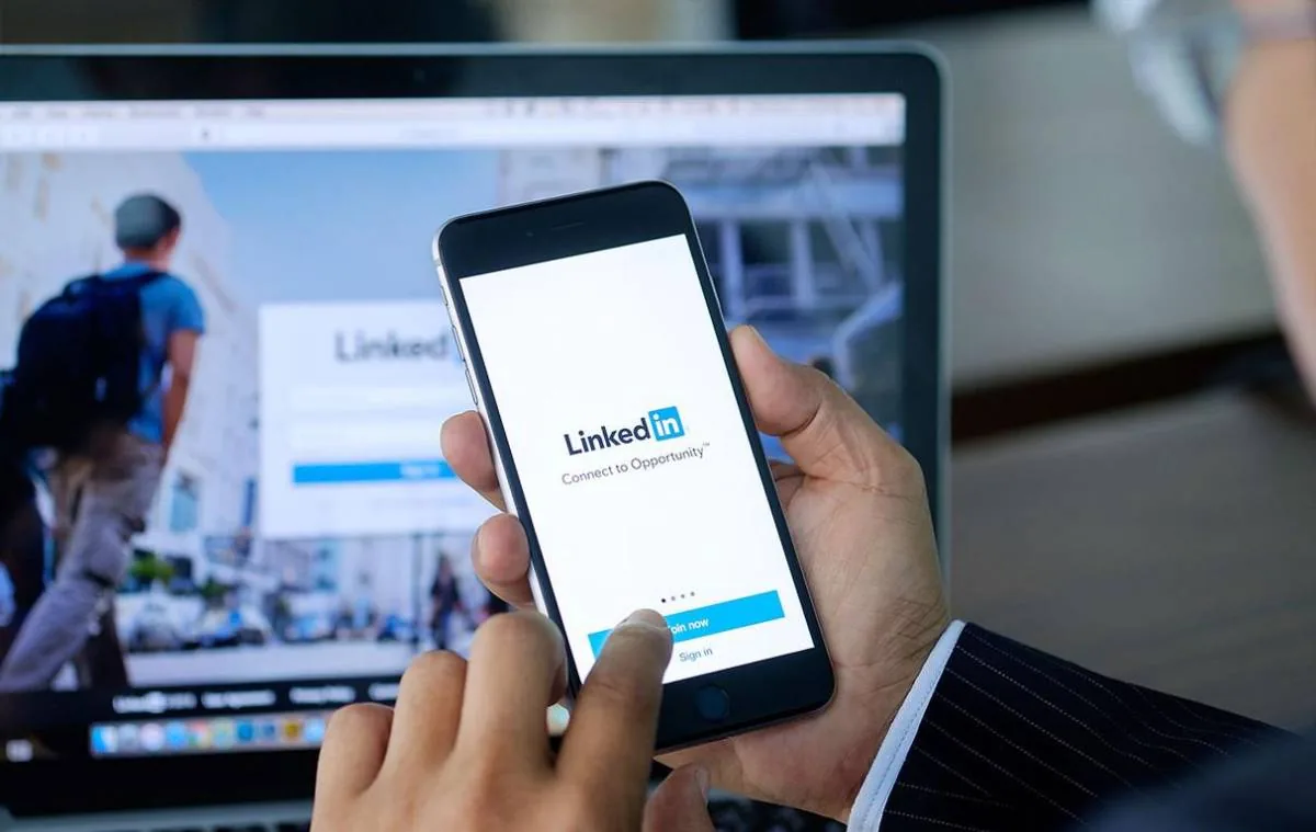 Una persona con traje sostiene un smartphone mostrando la app de LinkedIn, con el logo y 'Connect to Opportunity'. Un portátil en el fondo también muestra la web de LinkedIn.