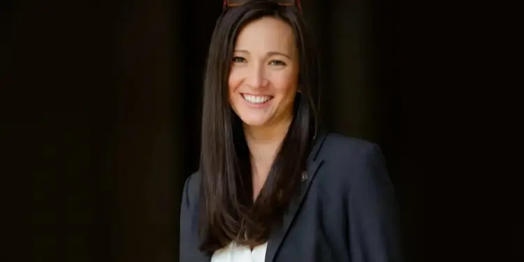 Retrato de mujer profesional sonriente con cabello oscuro y gafas en la cabeza, vestida con blazer oscuro y camisa blanca, sobre fondo oscuro.