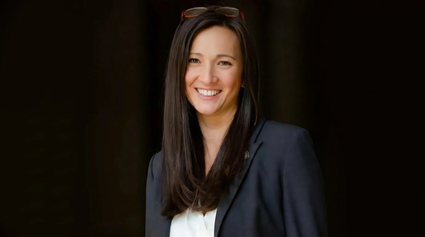 Retrato de mujer profesional sonriente con cabello oscuro y gafas en la cabeza, vestida con blazer oscuro y camisa blanca, sobre fondo oscuro.