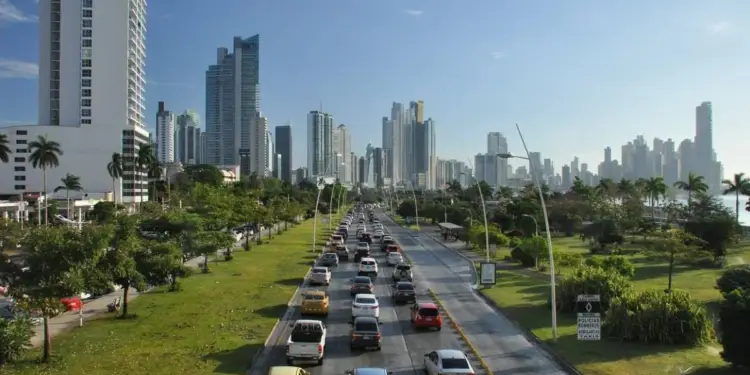 Vista elevada de una avenida urbana con congestión vehicular, rodeada de modernos edificios altos y zonas verdes.