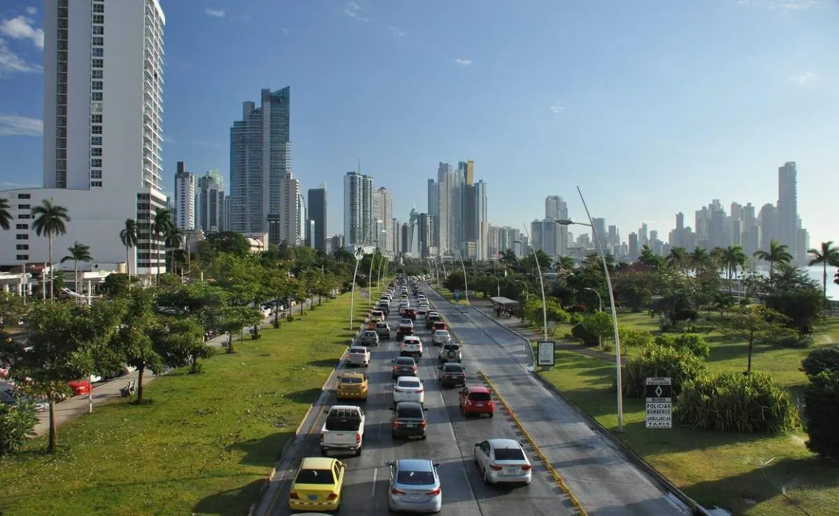 Vista elevada de una avenida urbana con congestión vehicular, rodeada de modernos edificios altos y zonas verdes.