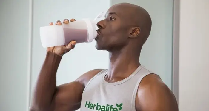 Hombre calvo vistiendo camiseta gris con la marca Herbalife bebiendo un batido de un vaso mezclador blanco.
