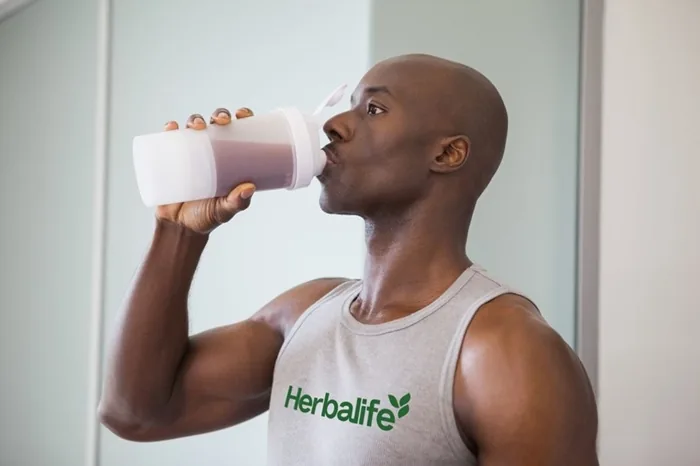 Hombre calvo vistiendo camiseta gris con la marca Herbalife bebiendo un batido de un vaso mezclador blanco.
