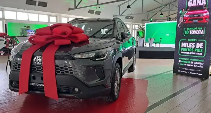 Automóvil gris oscuro con un gran lazo rojo en el cofre, exhibido en un salón amplio con piso de baldosas.