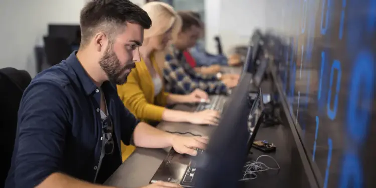 Un grupo de profesionales, incluyendo un hombre barbudo en primer plano, trabajando en laptops en un entorno de oficina moderno. Una pantalla grande con números digitales azules se ve al fondo, sugiriendo un enfoque en tecnología y datos.