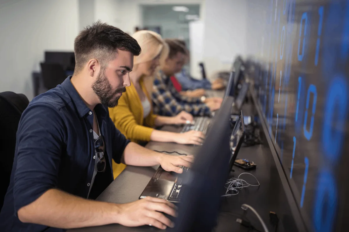 Un grupo de profesionales, incluyendo un hombre barbudo en primer plano, trabajando en laptops en un entorno de oficina moderno. Una pantalla grande con números digitales azules se ve al fondo, sugiriendo un enfoque en tecnología y datos.
