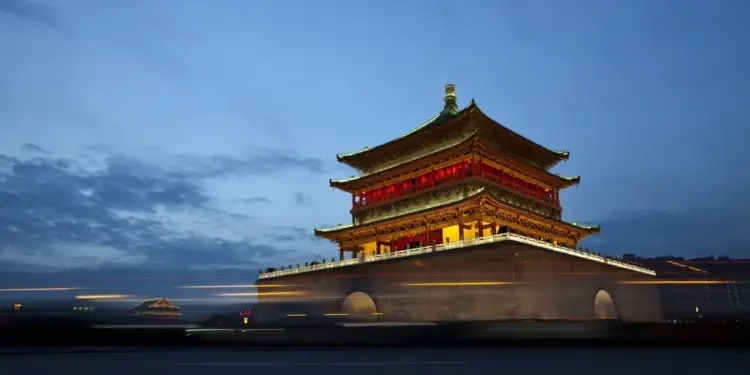 Imponente edificio tradicional asiático de varios niveles, iluminado con luces cálidas y rojas bajo un cielo azul oscuro al anochecer. Estelas de luz de vehículos cruzan el primer plano.