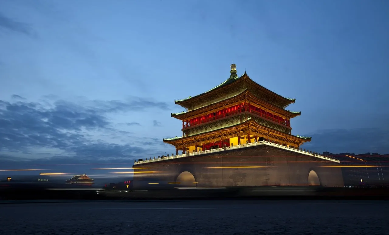 Imponente edificio tradicional asiático de varios niveles, iluminado con luces cálidas y rojas bajo un cielo azul oscuro al anochecer. Estelas de luz de vehículos cruzan el primer plano.