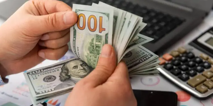 Manos contando un fajo de billetes de cien dólares sobre un escritorio con calculadora y computadora portátil.