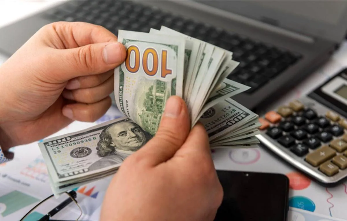 Manos contando un fajo de billetes de cien dólares sobre un escritorio con calculadora y computadora portátil.