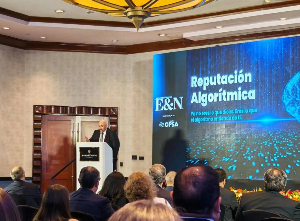 Hombre en traje dando una conferencia en el Hotel Intercontinental Guatemala sobre reputación algorítmica ante una audiencia. Pantalla muestra texto y logo E&N.